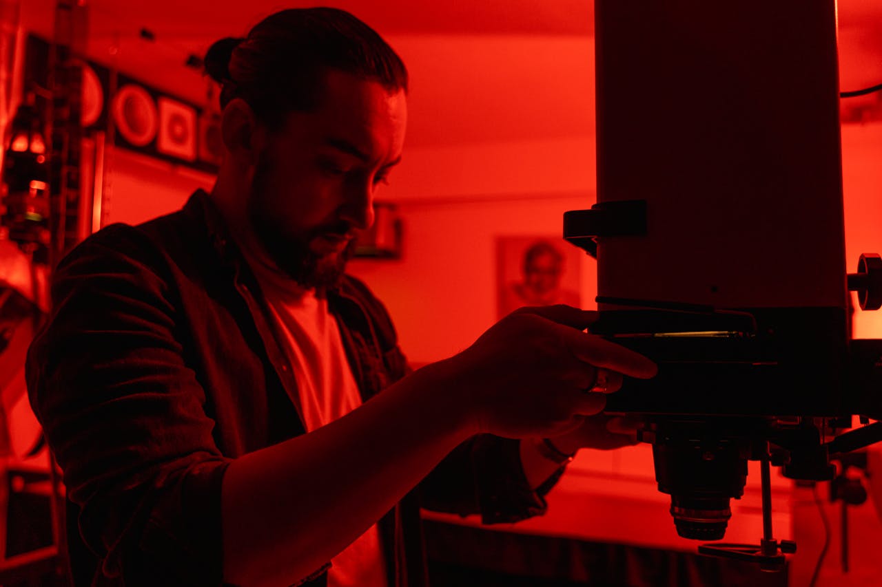 Photographer using an enlarger in a film photography darkroom with red lighting.